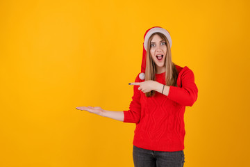 excited woman pointing at copy space on hand wearing santa hat and red sweater isolated over yellow