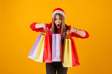 excited woman with gifts in holiday shopping bags wearing santa hat and sweater isolated over yellow
