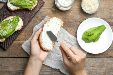Woman spreading cream cheese onto bread for avocado sandwich at wooden table, top view