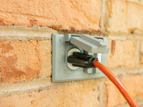 Power Cord Plugged Into Outdoor Electrical Wall Socket. Dual Covered Electricity Power Outlets On Brick Wall. Orange Extension Cord Plug Inserted Into Exterior Wall Jack.