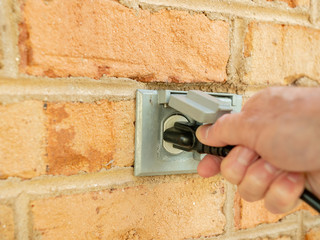 Man plugging power cord into outdoor electrical outlet. Dual covered power sockets on brick wall....