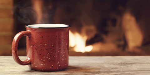 Red mug with hot tea in front of a burning fireplace, comfort, winter holidays and warmth of the...