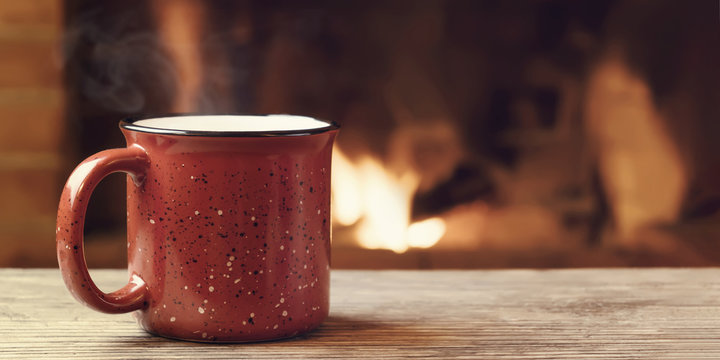 Red mug with hot tea in front of a burning fireplace, comfort, winter holidays and warmth of the hearth concept