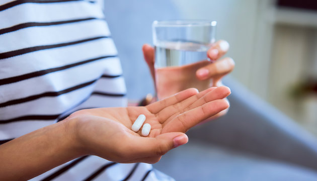 Woman holding white pill on hand and drinking water in glass on sofa in house, feels like sick. Health care concept.