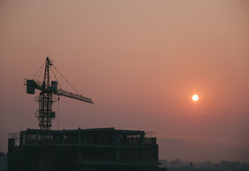 Crane building construction site at sunset or sunrise. Stock photo image silhouette of construction tower crane in sunset sky background and the sun. Building construction with crane during sunset