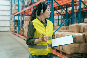 Elegant manager woman talking into headset in large warehouse. beautiful lady staff in safety vest walking in storehouse finding goods and parcels in shelves. girl employee making note on clipboard.