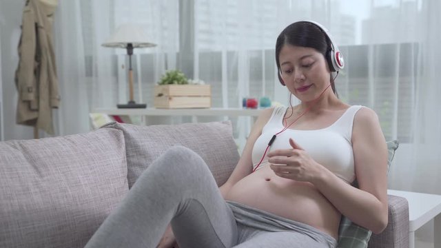 pregnancy motherhood technology people and expectation concept. happy pregnant woman with smartphone and headphones listen to music at home. young future mother touch bare belly while lying on sofa