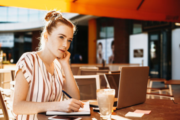 Charming young business woman with red hair and freckles taking notes while drinking coffee in a coffee shop.