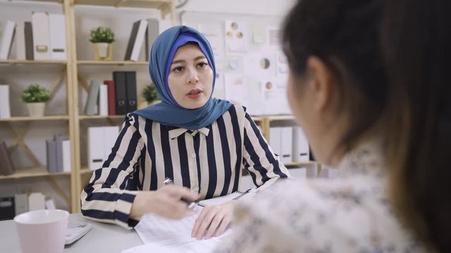 elegant muslim woman insurance agent wear blue hijab traditional sitting at workplace desk talking introducing project plan to chinese female customer. beautiful islam sales tryhard convince investor