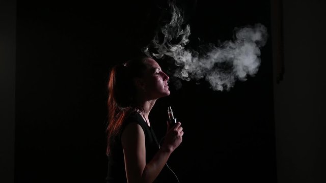 The brunette smokes an electronic cigarette on a black background, releases thick smoke from her mouth. Portrait of a woman smoking a vape, hovering.