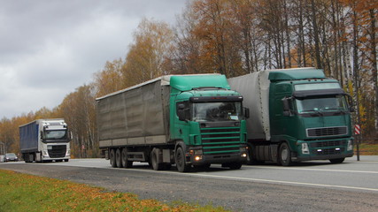 Fototapeta na wymiar Three European green semi trucks with awning trailers overtaking on asphalt country highway on autumn day, front side view, transport logistics, goods delivery, beautiful road landscape