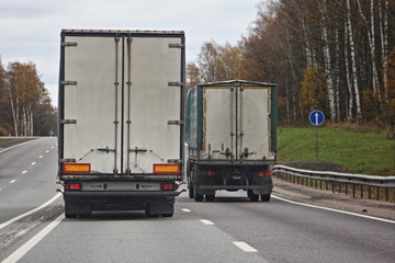 Logistics industry, international freight by road, two white trucks drive on all two-lane asphalted country road in autumn day, rear view, overtaking rules for drivers