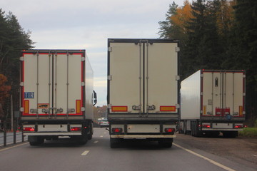 Three European white semi trucks with semi trailers overtaking on asphalt country road on autumn day, rear view, transport logistics, goods delivery