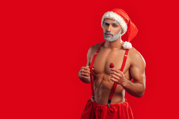 Christmas Freestyle. Young Santa Claus bare muscular upper body in hat standing isolated on red with strawberry posing confident copy space