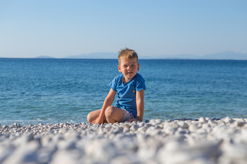 Fototapeta na wymiar Cute European little boy in a blue swimming T-short is standing in the sea. He is enjoying his summer holidays. Back view.