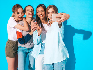 Four young smiling hipster women in summer clothes.Girls taking selfie self portrait photos on smartphone.Models posing near blue wall in studio,Female showing positive face emotions