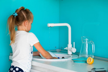 Little girl washes dishes at the kitchen sink in the kitchen