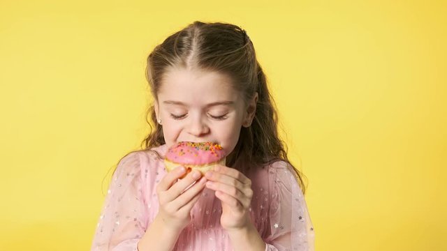 Little pretty girl eating donut on yellow background. Close-up portrait of cute caucasian girl in pink dress with pleasure eats doughnut with pink glase. Enjoying taste. 4K, UHD