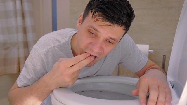 Portrait of man is vomiting in toilet sitting on the floor with two fingers in mouth, side view. Food poisoning and vomiting, cold and flu with high temperature. Sickness man at home with nausea.