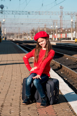 Portrait of a cute beautiful joyful girl sitting on a suitcase and waiting for a train at the train station