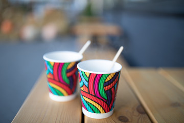 Yellow and red disposable paper cups with coffee close up against the light wood.