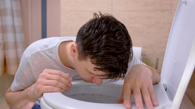 Man vomit in toilet sitting on floor looking at camera feeling bad having flu. Hangover, alcoholism, food poisoning and vomiting, cold and flu with high temperature. Sickness man at home with nausea.