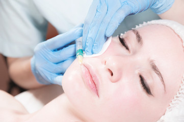 Obraz na płótnie Canvas Close-up Attractive young woman gets anti-aging face injections. She lies calmly in a clinic or salon. An experienced young cosmetologist fills female wrinkles with hyaluronic acid from a syringe