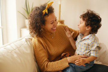Indoor shot of happy young Hispanic woman with brown wavy hair relaxing at home embracing her adorable toddler son. Cheerful mother bonding with infant son, sitting on sofa in living room, laughing - Powered by Adobe