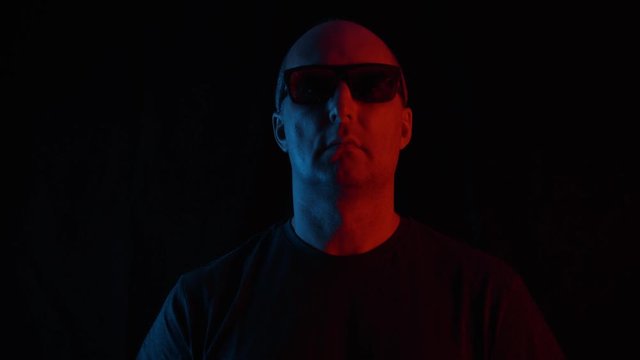 Serious middle aged man in sunglasses standing in darkness. Portrait of mature bald man in dark t-shirt and sunglasses standing and looking at camera on black background