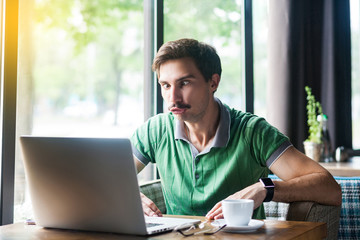 Young funny businessman in green t-shirt sitting and looking at laptop display with stupid crazy face and crossed eyes. business and freelancing concept. indoor shot near big window at daytime.