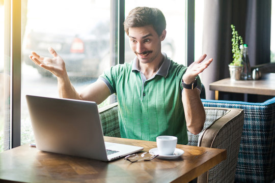 Young happy businessman in green t-shirt sitting and looking at laptop display on video call with raised arms and toothy smile. business and freelancing concept. indoor shot near big window at daytime