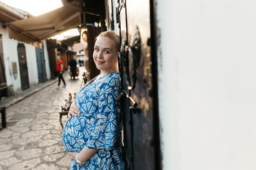 Portrait of beautiful pregnant woman. She is standing in the middle of the street and holding hands on her belly. 