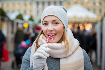 Young woman in Christmas market eating candy cane wearing knitted warm hat and scarf. Illuminated and decorated fair kiosks, shops and carousel on background. Helsinki, Finland