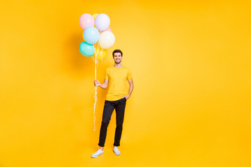 Full length photo of handsome guy holding many air balloons meeting guests at summer students party wear casual t-shirt black trousers isolated yellow color background © deagreez
