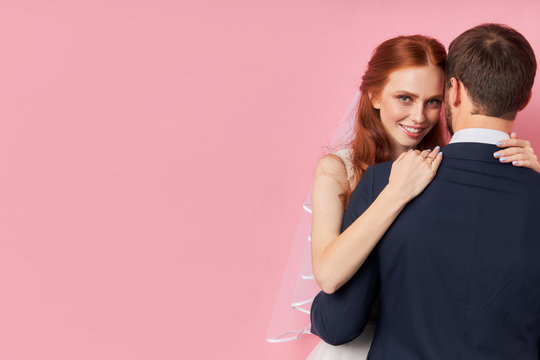 Back view on man in suit with her beautiful bride hugging him. Portrait of lovely couple isolated over pink background. Love concept