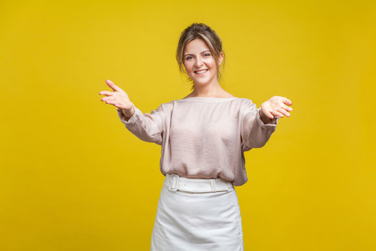Portrait of happy generous young woman with fair hair in casual blouse standing with raised hands and looking at camera, welcoming or giving smth. indoor studio shot isolated on yellow background
