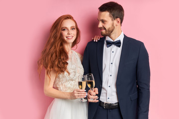 Caucasian nice-looking lovely cheerful couple celebrating their marriage drinking champagne isolated over pink background. Love, celebration concept. Man in tuxedo look with love at woman in dress
