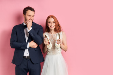 Fototapeta na wymiar Portrait of positive redhaired woman in suit seriously looking at smiling female in white wedding dress. isolated over pink background