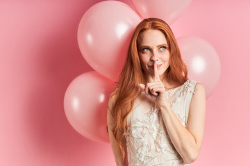 Obraz na płótnie Canvas Beautiful woman with auburn hair wearing white dress with air balloons behind, thinking, look up. Isolated over pink background