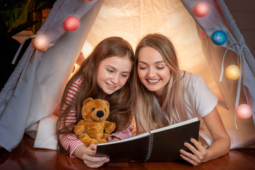 Happy mother and cute daughter reading book inside tepee tent together in bedroom, family relationship concept. © wirojsid