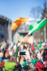 Female hands with mobile phone, making photo of traditional national Irish holiday party, carnival of St. Patrick's Day on a smartphone, selective focus.