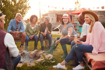 Group Of Mature Friends Sitting Around Fire And Drinking At Outdoor Campsite Bar - obrazy, fototapety, plakaty