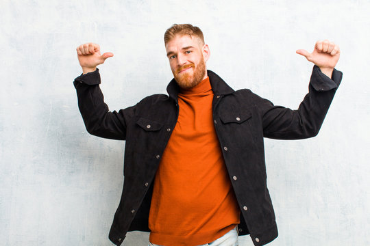 young red head man feeling proud, arrogant and confident, looking satisfied and successful, pointing to self against grunge wall
