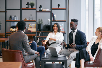 Cheerful business team sitting together on able discussing, smiling, break time, free time while working. Business meeting in office, african and caucasian partnership