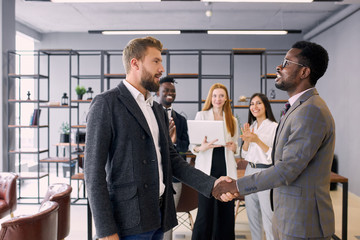 Two foreign executives in formal suits shaking hands in office while colleagues stand in background. Business people concept