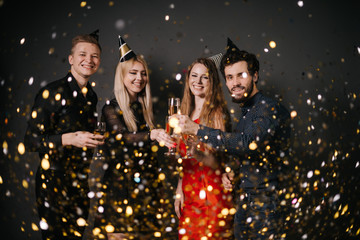 Obraz na płótnie Canvas Group of cheerful young people are clinking with champagne glasses. On friends from above the festive confetti falling. Shooting in professional studio on isolated black background.