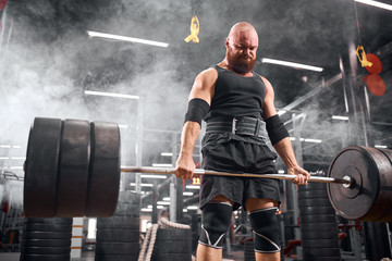 Strong active powerlifter, holding metal heavy barbell, trying to lift up, looking down, taking strength, standing in white smoke background, underground garage, shot from below