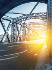 Asphalt road under the steel construction of a bridge in the city on a sunny day. Evening urban scene with the sunbeam in the tunnel. City life, transport and traffic concept.	