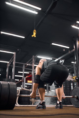Fototapeta na wymiar Bottom shot of strong brutal powerlifter dressed in black sportswear, bending in front of heavy barbell, standing with his back towards camera, modern fitness center concept, indoor shot