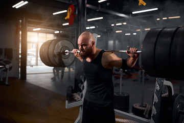 Strong active hairless athlete with thick brunette hard trying to rise hands with heavy barbell row above head, looking away with severe look, training in underground garage, black tone, portrait,